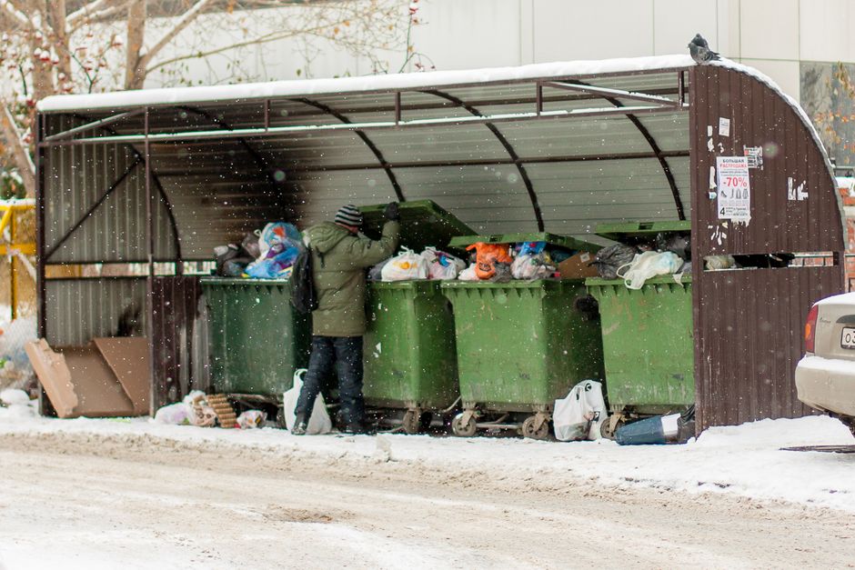 A person wearing a winter coat and striped beanie is seen sorting and disposing of various trash bags and cardboard boxes into large green waste bins inside a covered waste disposal area on a snowy day. The site is outdoors, with snow on the ground and snowflakes falling, positioned adjacent to a building and next to a parked car. The waste bins are lined up under a curved metal roof, which is brown on the sides and silver underneath. Around the waste area, there are additional cardboard pieces and miscellaneous rubbish lying on the snow, highlighting proper disposal in the context of home relocation or packing preparation, with the environment suggesting a routine collection process supported by local waste management and removal services such as Man with Van Leamouth.