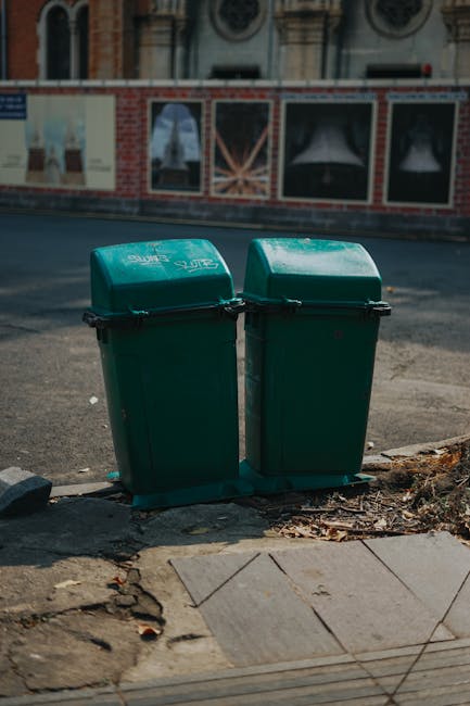 Two green wheelie bins with closed lids positioned side by side on a paved sidewalk adjacent to a curb, with some debris and dirt around their bases. Behind the bins, a section of asphalt road is visible, along with a building featuring large windows with black frames and decorative brickwork. The bins are outdoors in a street setting, suggesting a typical household waste disposal area as part of a home relocation or moving process. This scene reflects typical waste management practices encountered during removals or house moving activities, with Man with Van Leamouth providing services related to packing, transport, and logistics in the LEAMOUTH area.
