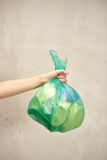 A person's arm holding a green plastic bag filled with waste materials, knotted at the top, against a plain, light-colored wall background. The bag appears to contain household rubbish, possibly including paper or plastic waste, and is used in the context of home clearances or rubbish collection services. The image illustrates waste disposal during a house removal or cleaning process, highlighting the importance of proper waste management for relocation projects. Occasioned by house removals, this scene reflects the task of loading waste into a bag for disposal as part of furniture transport and packing logistics coordinated by companies like Man with Van Leamouth, which offers removal services including bulky waste pickups in Leamouth.