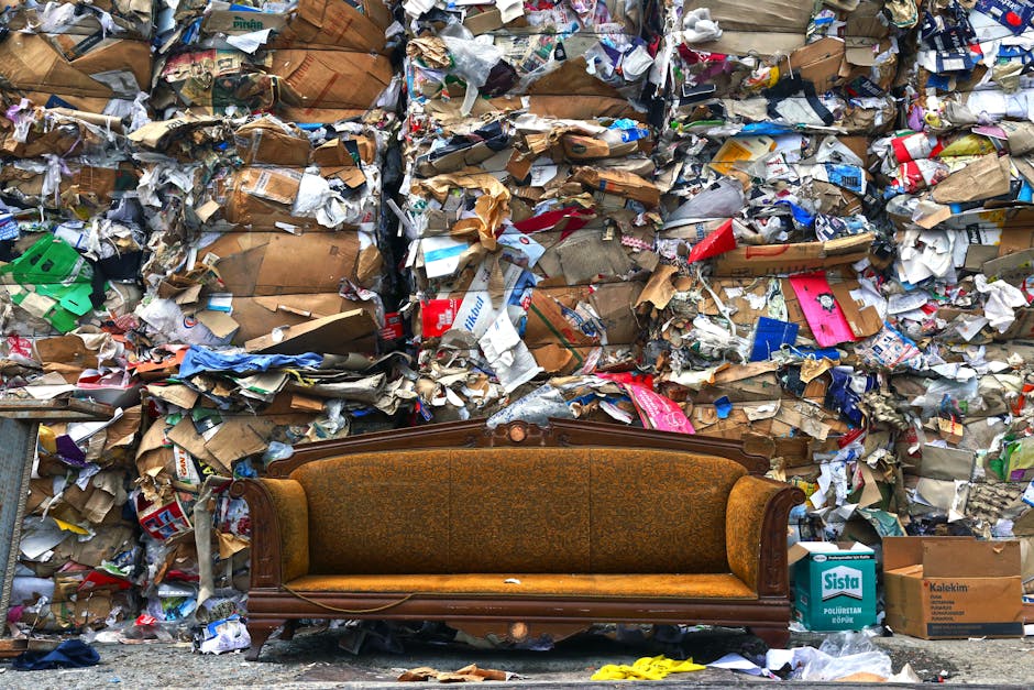 An old, upholstered wooden sofa with a patterned fabric seat and backrest is positioned in front of a large wall of tightly packed, mixed recyclable waste materials, including cardboard boxes, paper wrappings, plastic, and packaging debris, which are stacked high and appear to be awaiting collection or disposal. The setting appears to be outdoors, possibly on a pavement or loading area, with various boxes and packaging materials surrounding the sofa. This scene illustrates the logistics involved in bulk waste pickup and disposal, relevant to house removals and moving services offered by Man with Van Leamouth, highlighting the importance of proper waste management during home relocation projects.
