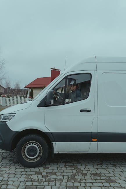 A man wearing a black cap, white t-shirt, and jeans is seen outdoors loading a large cardboard box into the side of a white panel van belonging to Man with Van Leamouth, parked on a residential street