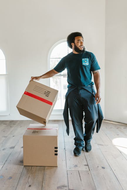 A man with dark curly hair, dressed in a blue t-shirt and black pants, is kneeling on a wooden floor inside a room filled with arranged cardboard moving boxes of various sizes, some labeled with dimensions such as 'Large 4.5 CU. FT.' and 'Medium 3.0 CU. FT.' He is holding a clipboard and appears to be inspecting or taking notes related to the packing and moving process. The boxes are stacked on the floor and some are positioned near a plain, light-colored wall with a slightly textured surface. There is natural lighting illuminating the scene, and a glimpse of a green plant can be seen on the right side of the image, suggesting an indoor environment prepared for the home relocation or furniture transport services provided by Man with Van Leamouth. The setting reflects a typical packing and loading phase during a house removals task, emphasizing careful documentation and organisation during the moving process.