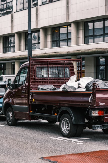 A burgundy commercial van parked on the side of a city street in front of a modern office building with large glass windows. The van's flatbed tray is loaded with various household items prepared for