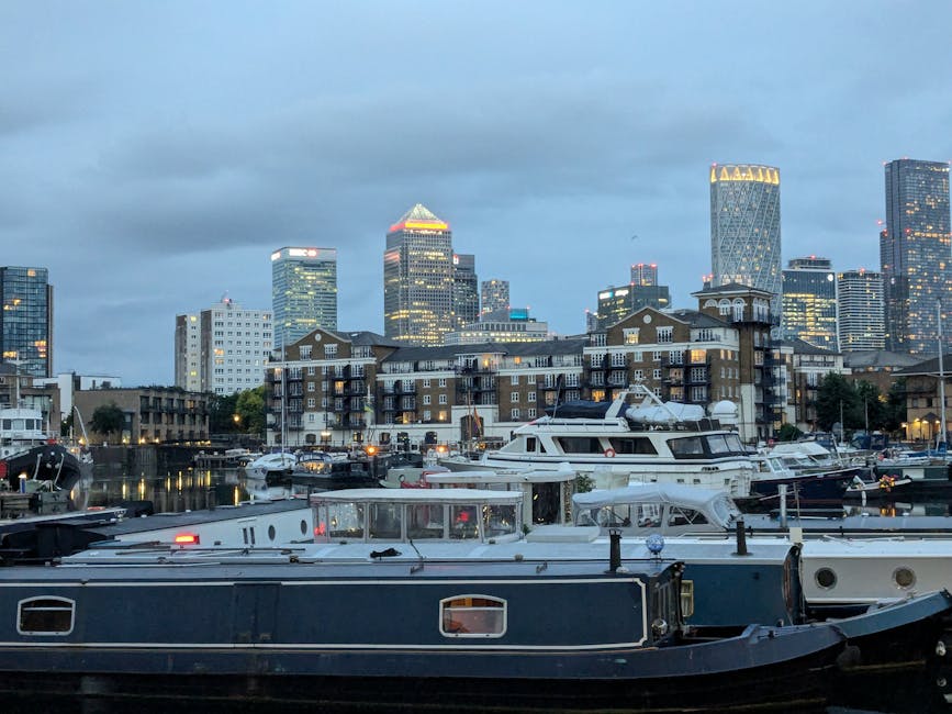 A view of a marina at dusk with several large boats and yachts docked along the water, some featuring visible cabin windows and deck structures. Behind the marina, there are residential buildings with