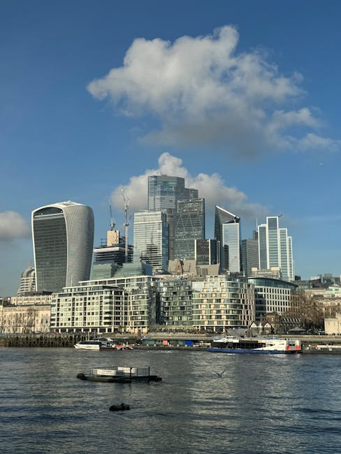 A panoramic view of London's city skyline featuring modern skyscrapers, including the distinct-shaped Walkie Talkie building and other high-rise office towers, set against a partly cloudy blue sky. In the foreground, the River Thames is visible with several boats and barges moored along the waterfront. The scene captures a typical day with natural light illuminating the buildings, which are part of the financial district near London City Island and Leamouth. The image is related to house removals and moving logistics, illustrating the urban environment often involved in home relocation and furniture transport services provided by Man with Van Leamouth, showcasing the setting where professional removals are conducted amidst iconic London architecture.