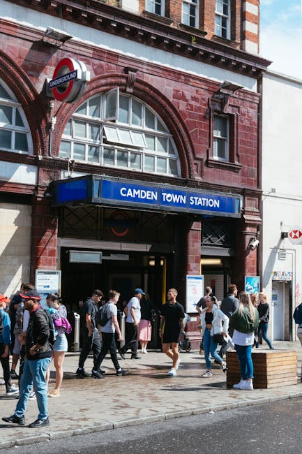 Exterior view of Camden Town Station with a blue and white sign displaying the station name, set in a red-brick building featuring arched windows and a rounded subway entrance. Numerous pedestrians ar