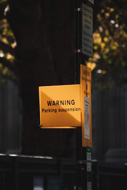 A close-up of a yellow warning sign attached to a metal pole on a pavement, displaying the message 'WARNING Parking suspension,' indicating temporary parking restrictions at Bow Creek. Behind the pole, a smaller white sign with additional parking information is visible, and part of a second yellow sign appears further up the pole. To the left, a large, dark tree trunk is partially visible, with blurred foliage and sunlight filtering through the leaves in the background. The scene suggests a street environment where parking may be restricted during a scheduled moving or home relocation process, which is relevant for planning furniture transport and loading logistics for house removals by Man with Van Leamouth. The lighting indicates late afternoon or early evening with natural daylight, emphasizing the importance of adhering to parking restrictions during a busy moving day.