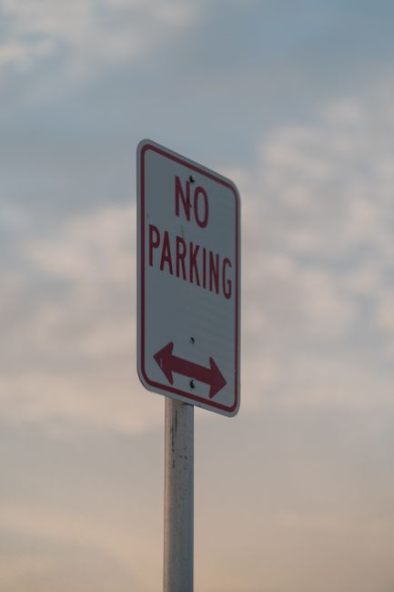 A rectangular 'No Parking' sign with a white background, red border, and bold red lettering, mounted on a metal pole situated on a pavement. The sign features an arrow pointing left and right at the bottom, indicating parking restrictions apply in both directions. In the background, the sky is visible with scattered clouds, suggesting an outdoor urban environment. The scene relates to parking regulations that may affect house removals or furniture transport during a home relocation, and the sign's positioning is relevant for planning moving logistics with Man with Van Leamouth, a company specialising in removals services. The image emphasizes the importance of understanding parking restrictions, particularly near properties where loading and unloading of furniture and packing materials are involved in the moving process.