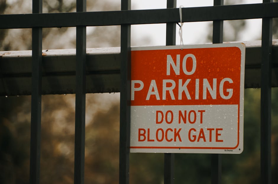 A close-up of a yellow warning sign attached to a metal pole on a pavement, displaying the message 'WARNING Parking suspension,' indicating temporary parking restrictions at Bow Creek. Behind the pole, a smaller white sign with additional parking information is visible, and part of a second yellow sign appears further up the pole. To the left, a large, dark tree trunk is partially visible, with blurred foliage and sunlight filtering through the leaves in the background. The scene suggests a street environment where parking may be restricted during a scheduled moving or home relocation process, which is relevant for planning furniture transport and loading logistics for house removals by Man with Van Leamouth. The lighting indicates late afternoon or early evening with natural daylight, emphasizing the importance of adhering to parking restrictions during a busy moving day.