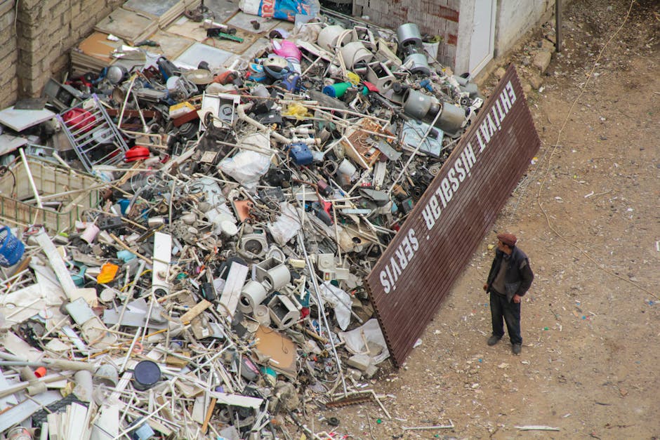 A man with curly black hair wearing a dark t-shirt and plaid shorts is standing outdoors next to a large, white open-top dumpster filled with various discarded building materials such as wooden planks