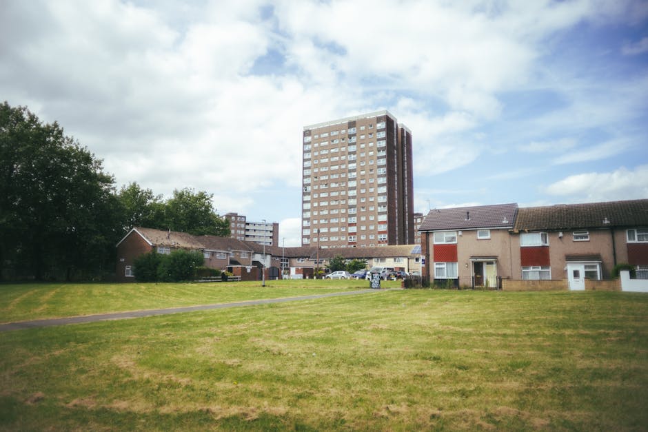 A large grassy open space in an urban residential area under a partly cloudy sky, with a paved pathway running through the foreground. In the background, there are multiple small, single-story houses with brick and stucco exteriors, some with red and white window shutters, and a taller multi-storey apartment building with a brown brick facade and numerous windows. Several parked cars are visible near the houses. The scene appears to be during daytime, with natural light illuminating the area. This setting reflects a typical environment where house removals and furniture transport might take place, and is relevant to the process of packing, loading, and transit involved in home relocation services carried out by companies like Man with Van Leamouth.