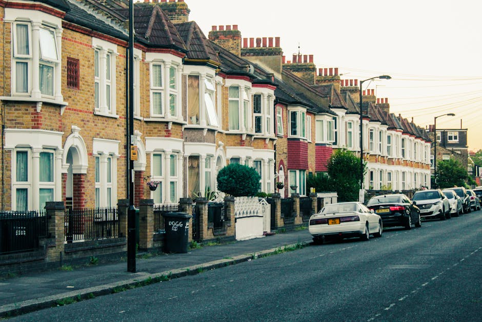 A row of traditional terraced houses in Leamouth, Tower Hamlets, with brick facades, bay windows, and decorative window frames, situated on a residential street during daylight. The pavement is lined with black metal railings, and a few small trees and bushes are visible in front of the houses. Several cars are parked along the street, including sedans and hatchbacks, and utility poles with overhead wires run parallel to the road. The scene captures the quiet atmosphere of a typical UK neighbourhood, and the image relates to home relocation and furniture transport processes, which [COMPANY_NAME], such as Man with Van Leamouth, facilitates through professional removals and packing services.