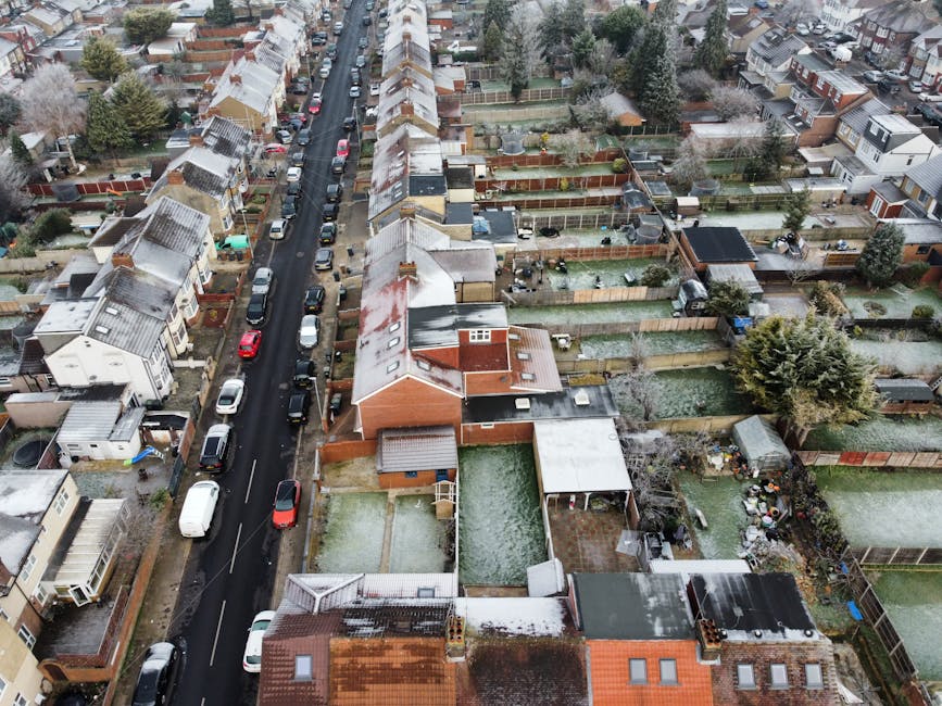A large grassy open space in an urban residential area under a partly cloudy sky, with a paved pathway running through the foreground. In the background, there are multiple small, single-story houses with brick and stucco exteriors, some with red and white window shutters, and a taller multi-storey apartment building with a brown brick facade and numerous windows. Several parked cars are visible near the houses. The scene appears to be during daytime, with natural light illuminating the area. This setting reflects a typical environment where house removals and furniture transport might take place, and is relevant to the process of packing, loading, and transit involved in home relocation services carried out by companies like Man with Van Leamouth.