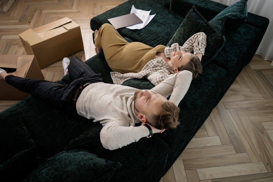A young woman and a young boy are lying on a dark green velvet sofa inside a living room, appearing relaxed and smiling while enjoying a moment together during a home relocation process. The woman is wearing a beige skirt and a checked shirt, with her head resting on a cushion and her hand behind her head. The boy, dressed in a light-colored sweater and dark plaid trousers, is lying beside her with his head turned towards her, both looking happy. Next to the sofa, on the wooden floor, there are cardboard moving boxes, some open and partially filled with packing materials, indicating recent or ongoing packing activities for a house move. The room is well-lit with natural light, and the scene is set on a herringbone-patterned wooden floor, suggesting a residential environment managed by a professional removals company such as Man with Van Leamouth, who focus on house removals and furniture transport during household relocations.