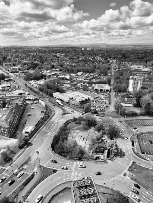 A black and white aerial photograph showing an extensive view of a town with a large roundabout at the centre, surrounded by multiple lanes of traffic with cars moving in different directions. Adjacent to the roundabout is a landscaped area with trees and grass, and nearby are commercial and residential buildings. On one side of the roundabout, a curved road leads to a parking lot filled with vehicles. In the background, there are clusters of trees, low-rise commercial structures, and a few taller buildings, with a broader landscape of fields and distant hills under a sky filled with scattered clouds. The scene captures the busy nature of the town's transportation infrastructure, which could be relevant to a home relocation or furniture transport process facilitated by Man with Van Leamouth during house removals and moving services.